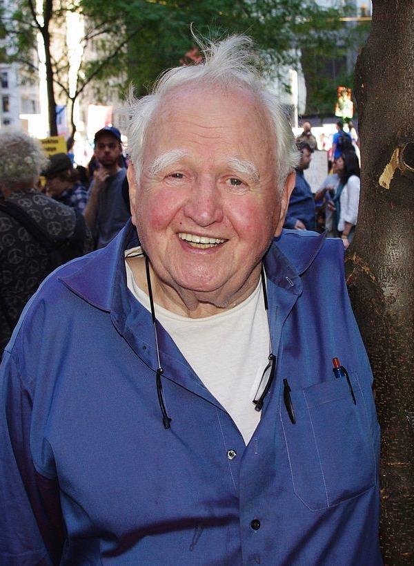 an older man with white hair and bushy eyebrows smiles at the camera outdoors at a public gathering