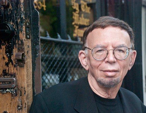 David Evanier, with wire-rim glasses, in black jacket & black T-shirt, outside, in front of a gate. 