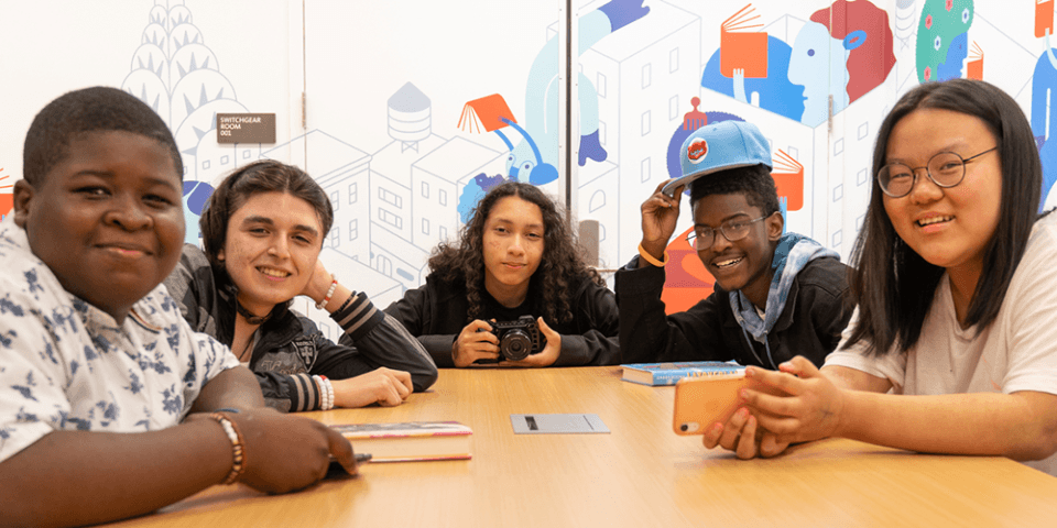 A diverse group of teens sits at a long wooden table, smiling at the camera; in the background is a colorful mural. 