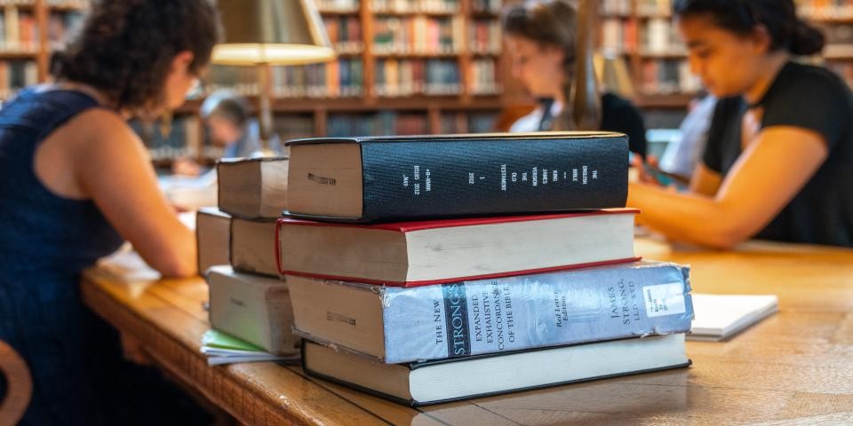 A large stack of books sits on a table; in the background, three researchers study.