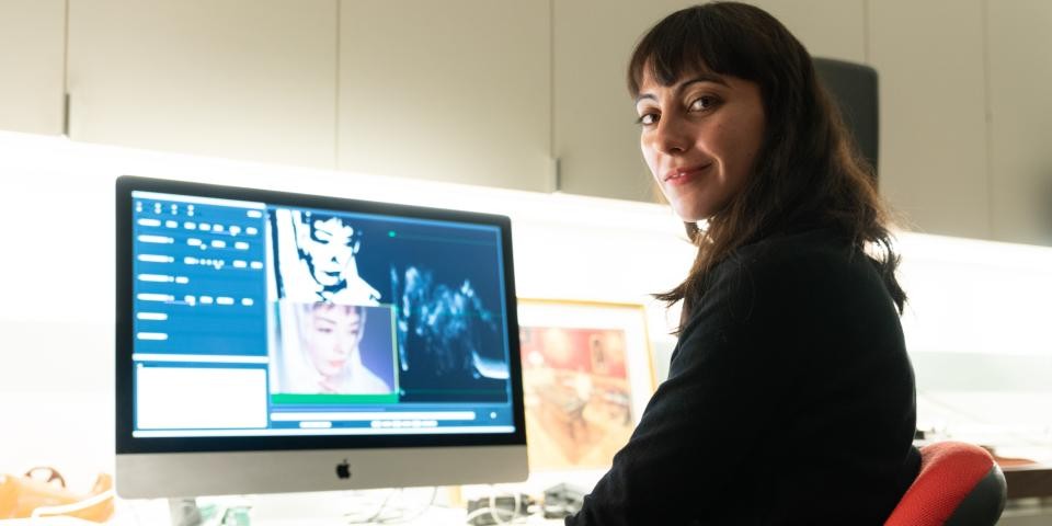 A woman sits a computer with film stills on the screen, she is looking back towards the camera and smiling.