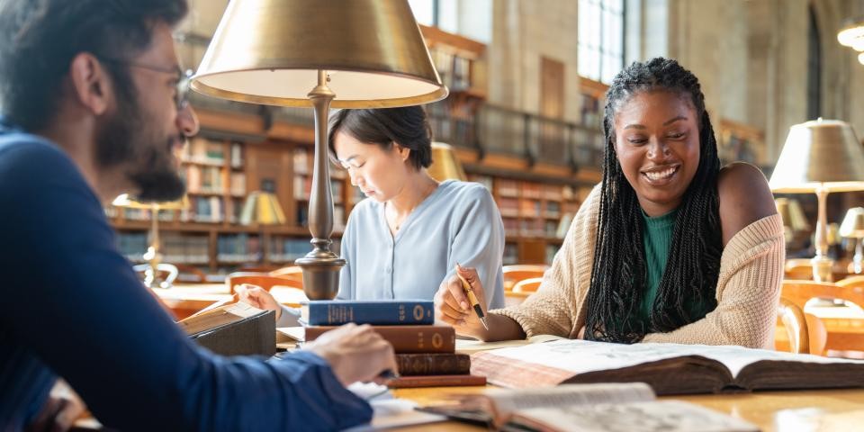 Three researchers sit at a table filled with books in the Rose Main Reading Room. 