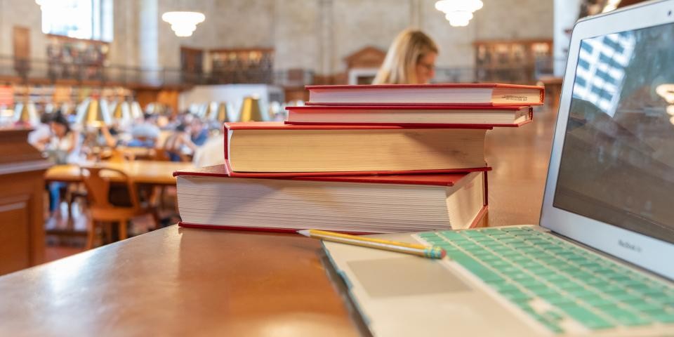 A laptop with green keys is in the foreground; behind it, a stack of books and a room full of researchers. 