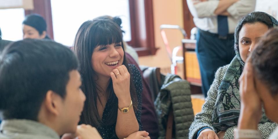 A group of people smile and chat in a brightly lit room with windows.