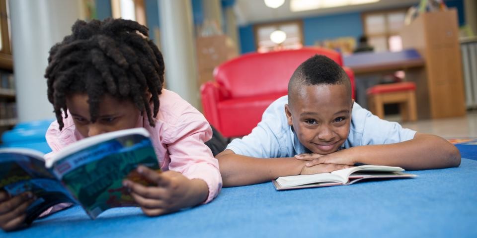 Two boys lay on the ground reading books, one of the boys smiles at the camera.