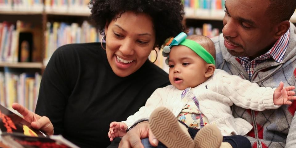 A woman and a man read a picture book to a small baby in a green ribbon.