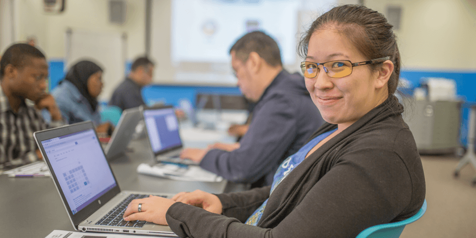 A woman sits at a table with a laptop and smiles at the camera; in the background, there are other laptop users. 