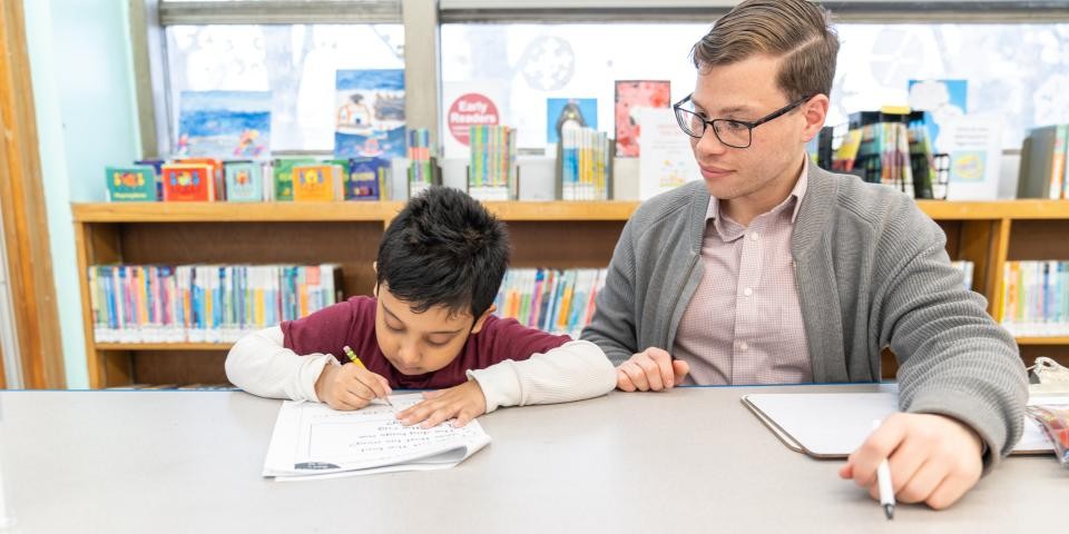 An adult looking and a child sitting down at a table in a library setting.