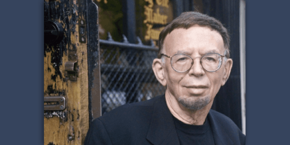 David Evanier, with wire-rim glasses, in black jacket & black T-shirt, outside, in front of a gate. 