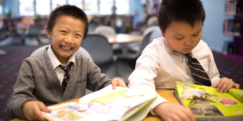 Two young boys read books while smiling and sitting at a table.