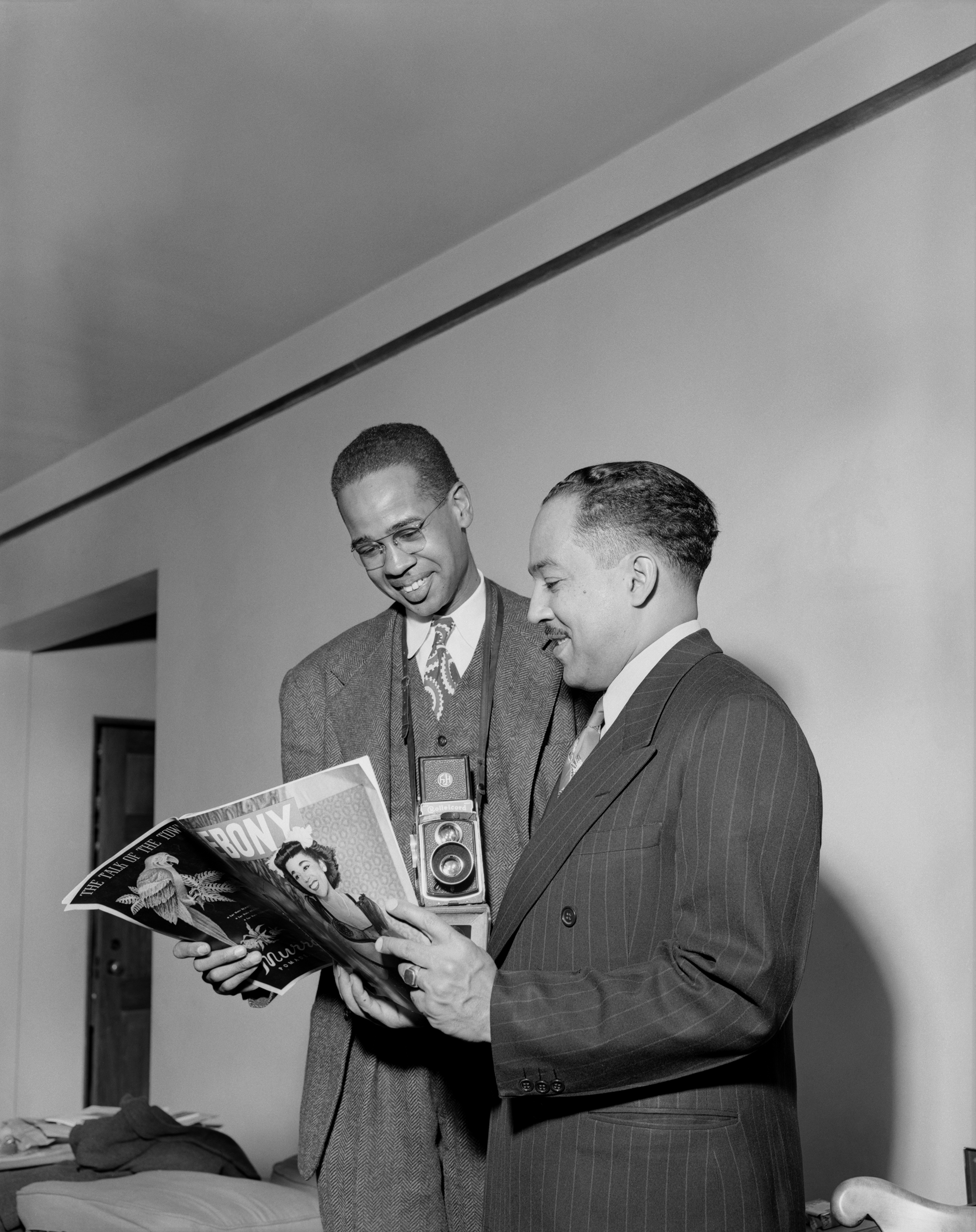 Image of two Black men standing, reading a magazine. The man on the left has a camera hanging around his neck.