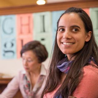 Photo of a young woman in the foreground wearing a pink shirt and smiling during a class at the library