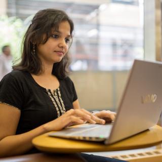 A library patron types on a laptop while at a small table in a brightly lit room. 