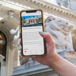 A hand with green nail polish holds a phone and headphones outside of the flagship NYPL building.