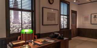 In an office on a large wooden desk sits a typewriter, rotary phone, desk blotter, accountant lamp, and other office supplies from the 1950s; The two office windows have projected scenes of the NYC Skyline featuring the Empire State Building.