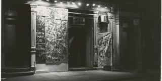 The front of a performance venue at night is pictured. The door is lighted by a row of stage lights. The windows are covvred are plastered in paper and every inch is covered in scrawled text. A draped fabric sign next to the door reads 'Caffe Cino'