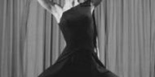 In a black and white photograph a dancer in draping black dress raises their arms