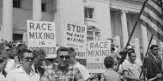 In a black and white photograph of a 1950s demonstration a group of white adults hold American flags and signs against integration. 