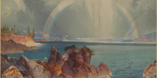 From the rocky shore of a large lake a rainbow is seen arcing over the middle of the lake. 