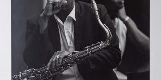 A black and white photograph of a Black man and woman. The man is in the foreground holding a saxophone in his lap with his right fist resting in front of his face. The woman is in the background in less clear focus