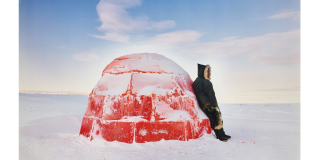 Man in winter coat leaning on a red igloo, in a snowy scenery 