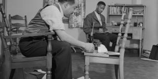 Black and white photo of Langston Hughes seated, working using a chair as a workstation, surrounded by papers on the floor and a man in the background also working