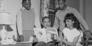 Black and white photo of Langston Hughes seated, reading, surrounded by three students, two young men standing and a young women seated beside Hughes