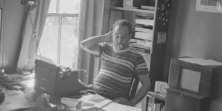 Black and white photo of Langston Hughes in a striped shirt with a cigarette dangling from his lips, sitting at a desk with a typewriter surrounded by papers.