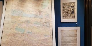 Photograph of a large map, an illustrated print, and a handwritten letter in a display case against a blue background
