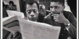 Black and white photo of James Baldwin seated beside Medgar Evers reading the newspaper