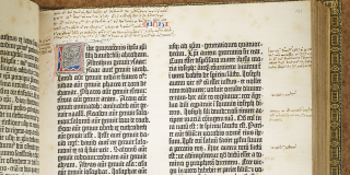 A Gutenberg Bible displayed open to show closely printed text in Latin in two columns, featuring an illuminated letter in the top left corner along with various handwritten annotations in the margins.