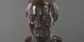 Bronze bust of Voltaire by Jean-Antoine Houdon; item is dark brown in color; Voltaire is balding with a very slight smile on his face