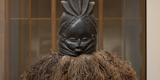 Helmet-like mask of dark wood carved in the round, with a face at the front, and surrounded by a skirt of yellow and brown raffia strands