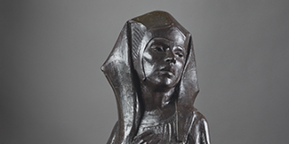 A dark bronze sculpture of a figure wearing a headpiece looking longingly over their left shoulder with their right hand over their heart 