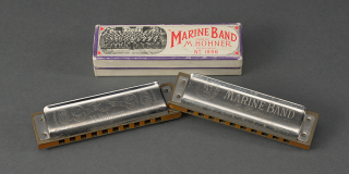 Two silver harmonicas are displayed next to each other at different angles, with the left edge of the harmonica on the right stacked on top of the right edge of the harmonica on the left. A harmonica box is placed behind them. The instruments and the box all bear the company name “Marine Band”