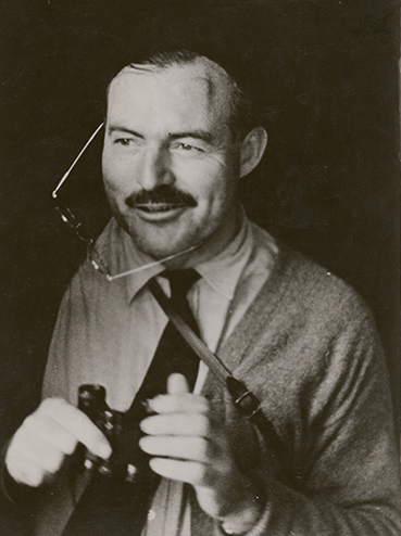 Historic black-and-white photo of Ernest Hemingway smirking and looking in the distance with a pair of glasses hanging from one ear while he holds a pair of binoculars.