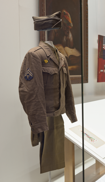 Three-quarter view of dark khaki uniform, with pants folded over hanger beneath a shirt and jacket on a form and hat displayed above