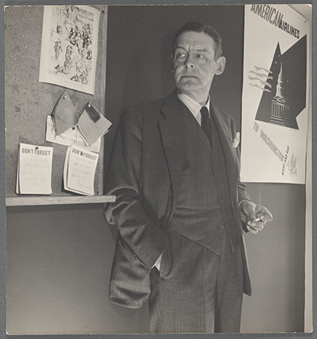 Black-and-white photograph of T.S. Eliot wearing a suit and tie, standing between a bulletin board and an American Airlines poster, holding a lit cigarette and looking off to his right