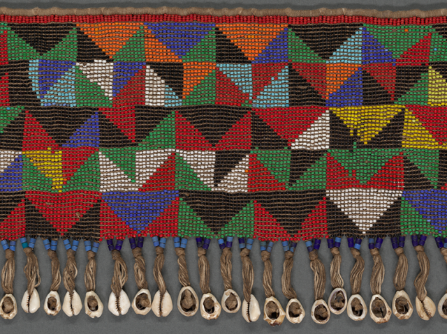 Beadwork in triangular and geometric patterns of many colors, attached to a threaded “belt” and decorated at bottom with a row of cowrie shells dangling from gathered threads