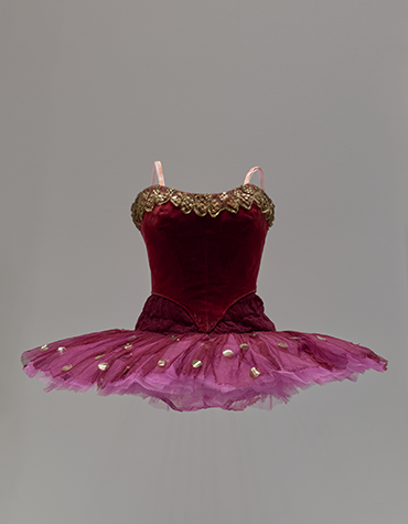 Photograph of a dress form displaying a red velvet ballet tutu decorated with sequins and gold trim 