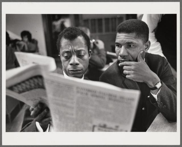 Black and white photo of James Baldwin seated beside Medgar Evers reading the newspaper