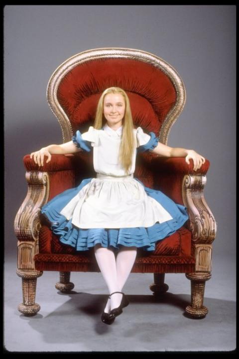 Actor Kate Burton is shown costumed as Alice in the trademark blue and white dress. Burnton smiles at the camera while sitting in a large ornate throne.