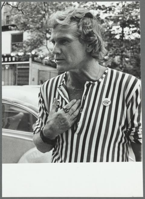 A man in a striped shirt looks away from the camera with a furrowed brow and a hand on his chest. 