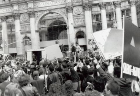 In a 1989 black and white photograph a large group of protestors is pictured outside of New York City Hall. Signs saying 'Silence=Death" can be seen throughout the crowd. 