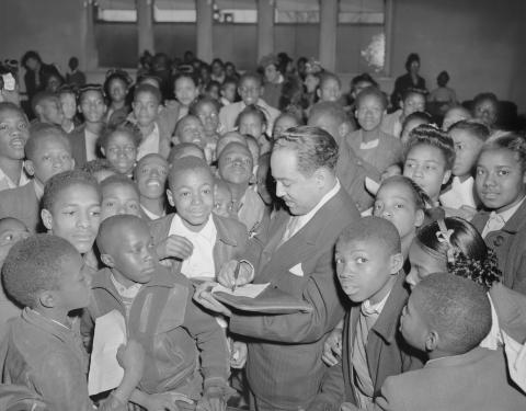 Black and white photo of Langston Hughes in a suit signing an autograph surrounded by a sea of black children