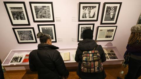 Color image of three people in an exhibition space looking down into a display case of books and letters. The wall in front of them is lavender with two quads of black and white photos.