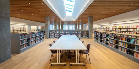 Interior of Inwood Library, featuring a large skylight, hardwood details, bookshelves, and a long table with chairs. 