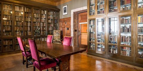 Photograph of an empty reading room interior with a wooden table, purple velvet chairs and stacks of books along the walls